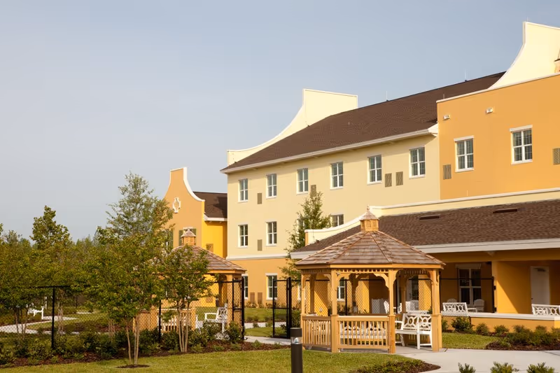 Yellow multi-story senior living building with a landscaped courtyard and wooden gazebos.