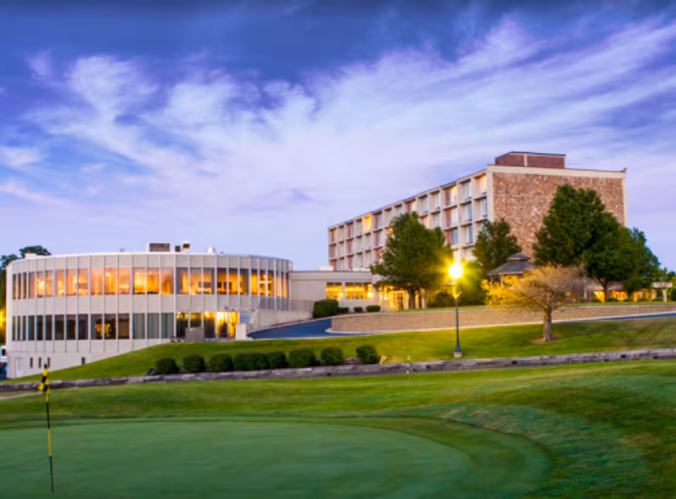 Exterior view of a multi-story senior living building with a round glass wing and a putting green in the foreground at dusk.