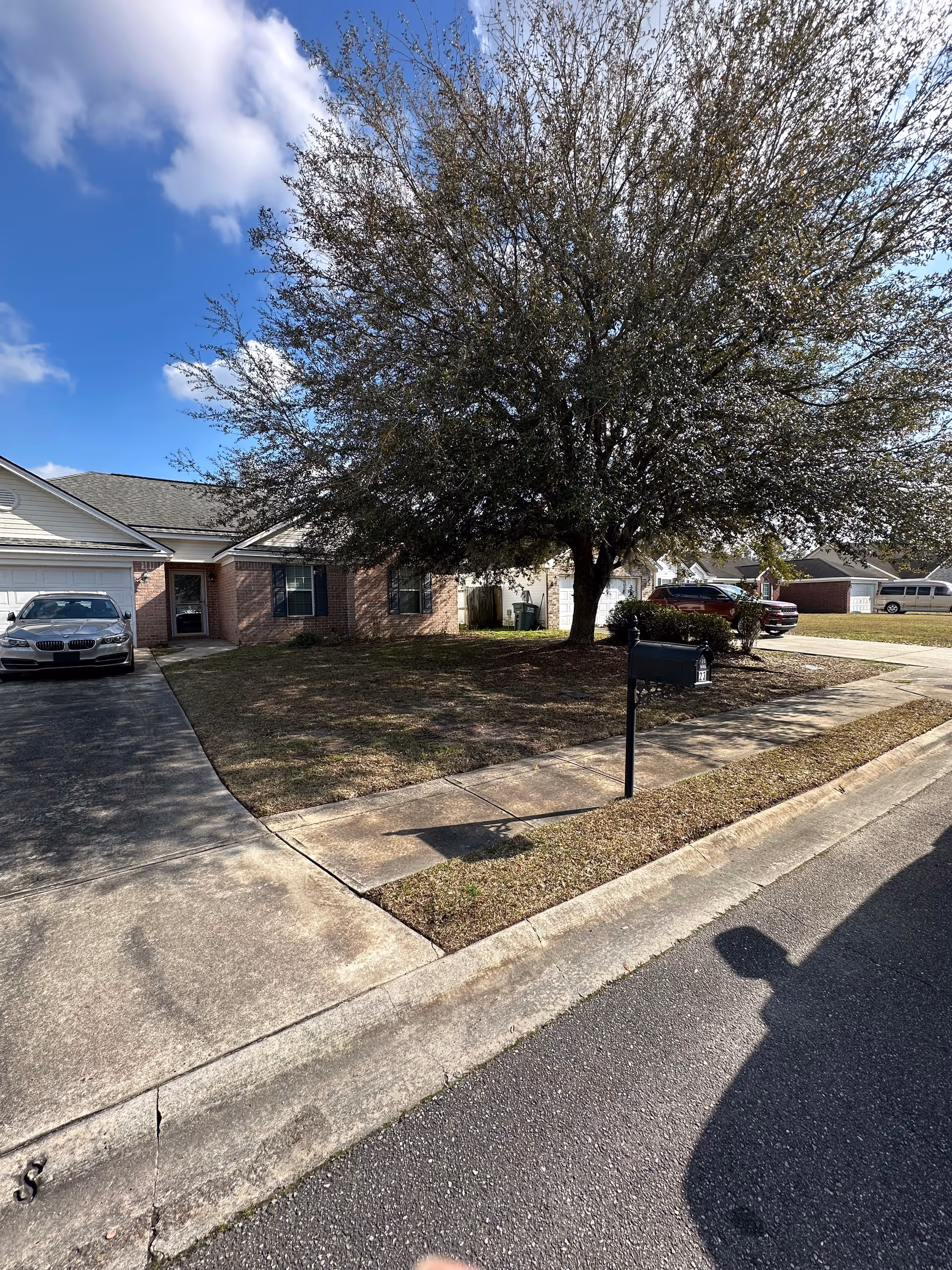 Suburban single-story brick home with a large tree, driveway, and parked cars.