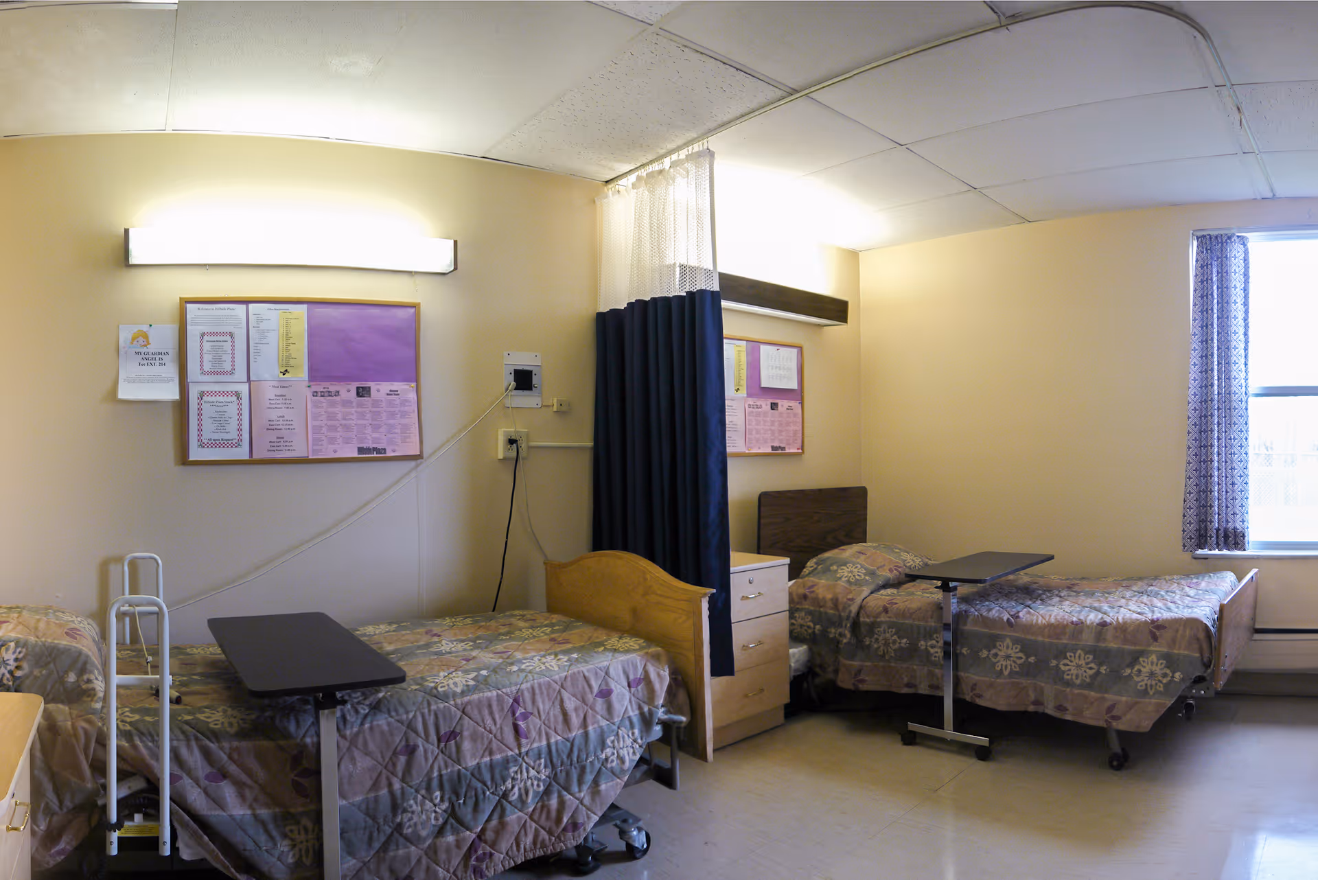 A shared bedroom in a senior living facility with two single beds, each with a floral patterned bedspread and an overbed table. There is a curtain divider between the beds, a window with patterned curtains, a bulletin board on the wall, and a walker next to one of the beds.