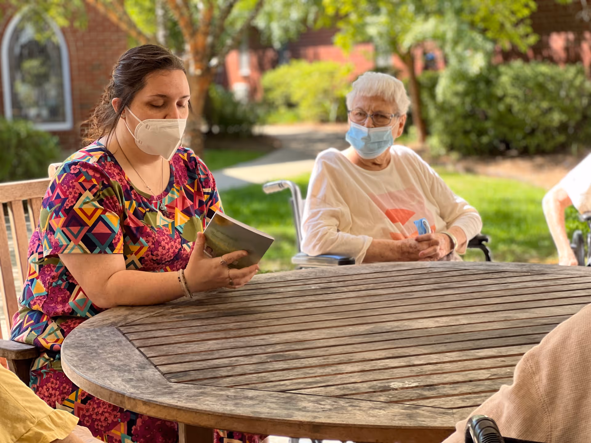A woman wearing a colorful patterned dress and a face mask is reading a book while sitting at a round wooden table outdoors. An elderly woman in a wheelchair, also wearing a face mask, is sitting nearby, listening attentively. The background shows greenery and a brick building.