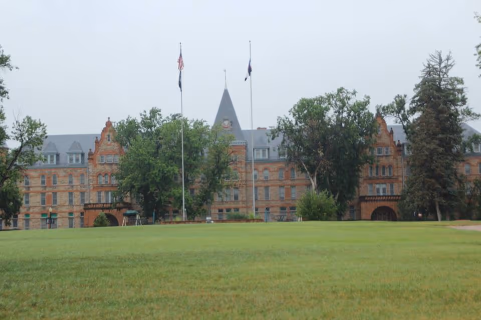 Wide view of a large historic brick institutional building with a central pointed tower, flagpoles, trees, and a grassy lawn in front.