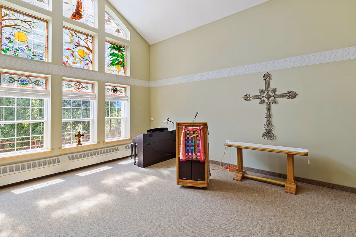 A bright room with large stained glass windows depicting colorful designs, a wooden podium with a colorful cloth, a black piano, a wooden bench, and a decorative metal cross mounted on the wall.