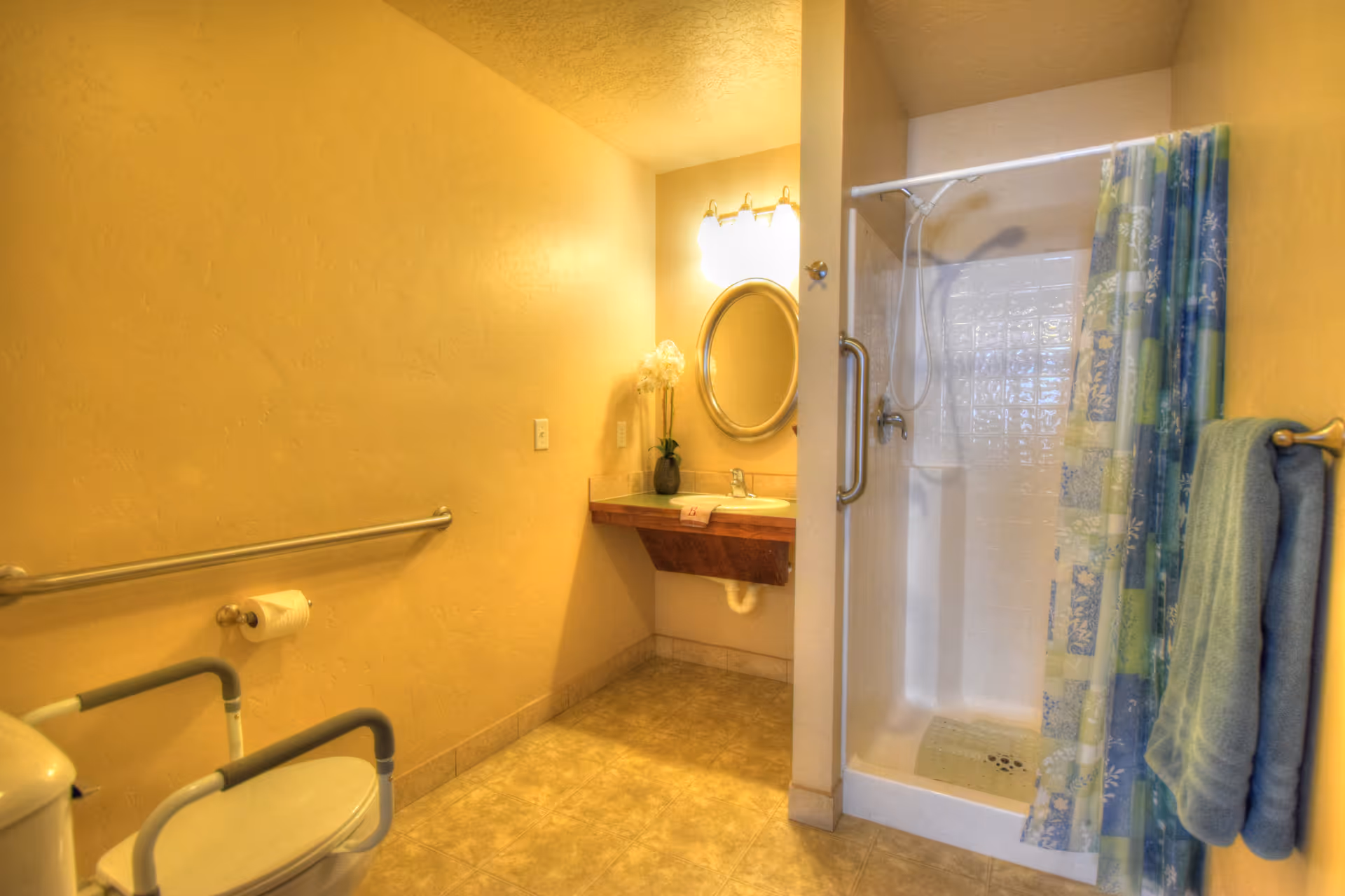 A bathroom with beige walls and tiled floor featuring a toilet with armrests, a grab bar, a toilet paper holder, a sink with a wooden countertop, an oval mirror above the sink, a vase with flowers, a shower with a grab bar and a colorful shower curtain, and towels hanging on a rack.