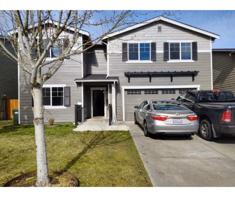 Two-story gray townhouse-style home with front door, attached garage, driveway, parked cars, and tree.