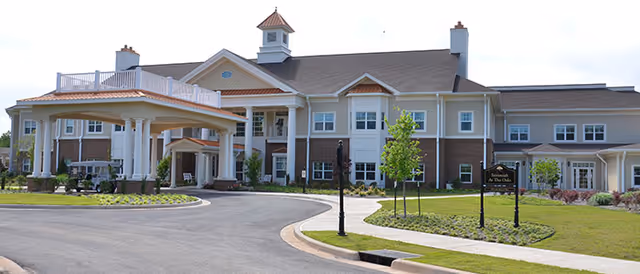 Exterior view of a large, two-story senior living facility building with a covered entrance, multiple windows, and a well-maintained lawn with small trees and shrubs.