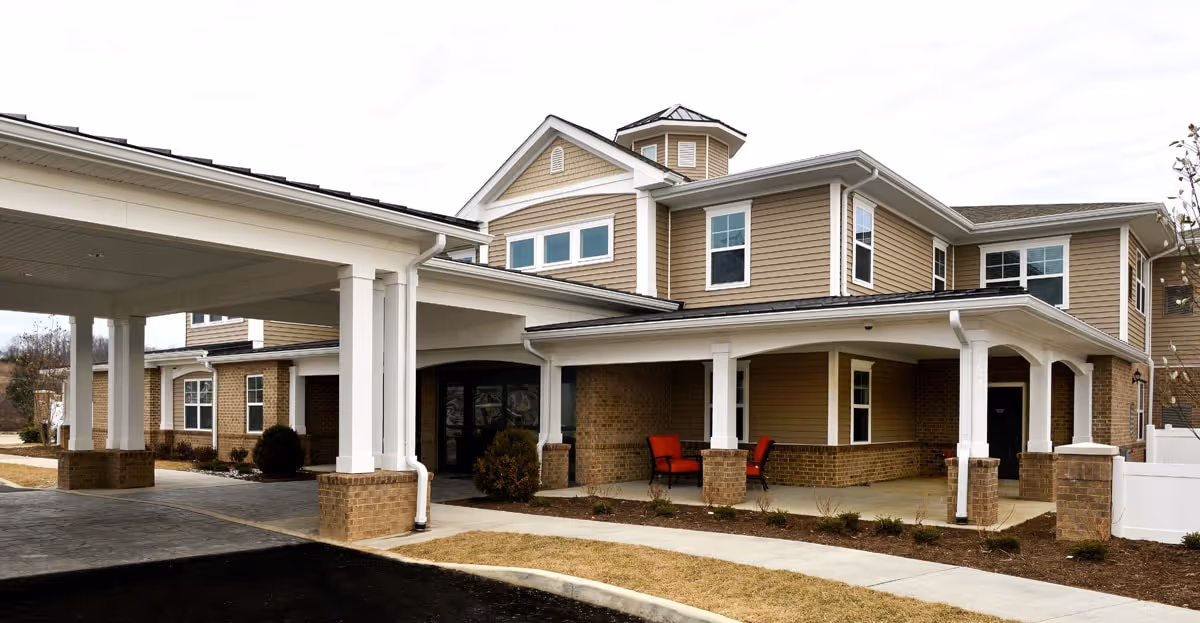Exterior view of Bellaire at Devonshire Assisted Living & Memory Care building with a covered entrance supported by white columns and brick bases. The building has beige siding with white trim and multiple windows. There are two red chairs placed under the covered porch area.