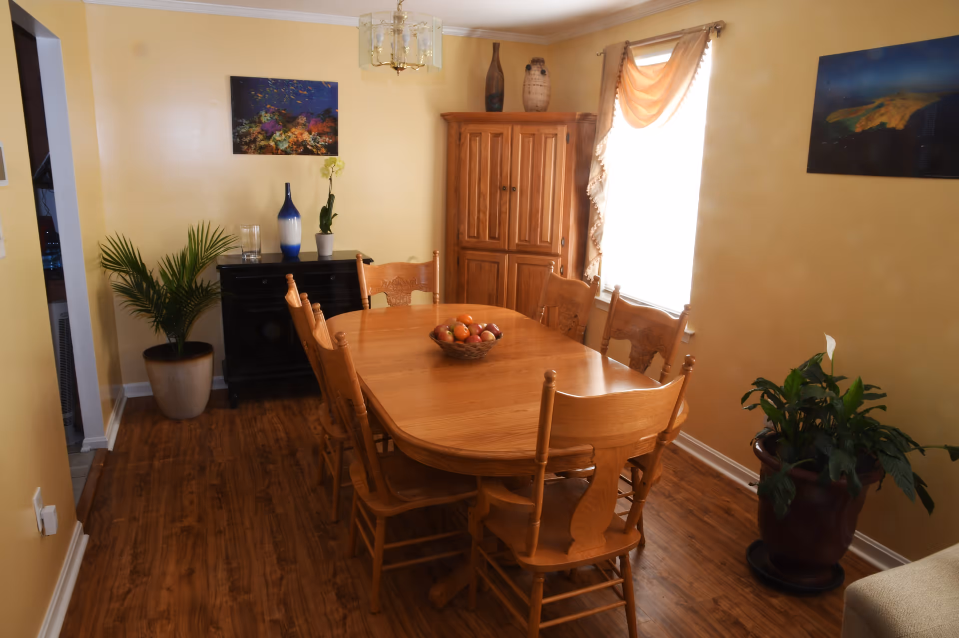 A dining room with a wooden dining table and six matching chairs. A bowl of fruit is placed in the center of the table. The room has yellow walls, wooden flooring, and two framed pictures hanging on the walls. There is a wooden cabinet in the corner, a black sideboard with decorative vases and a plant on top, and two potted plants on the floor. A window with a draped curtain allows natural light into the room.