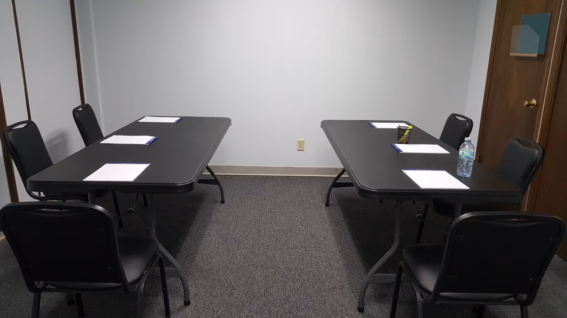 Two black tables with chairs in a small meeting room; papers and a water bottle present.