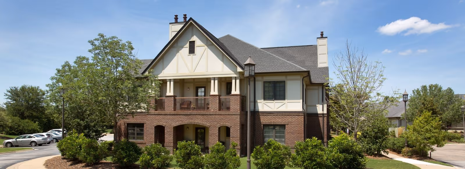 Two-story brick and stucco building with a covered second-floor balcony, landscaped shrubs, and parked cars under a blue sky.