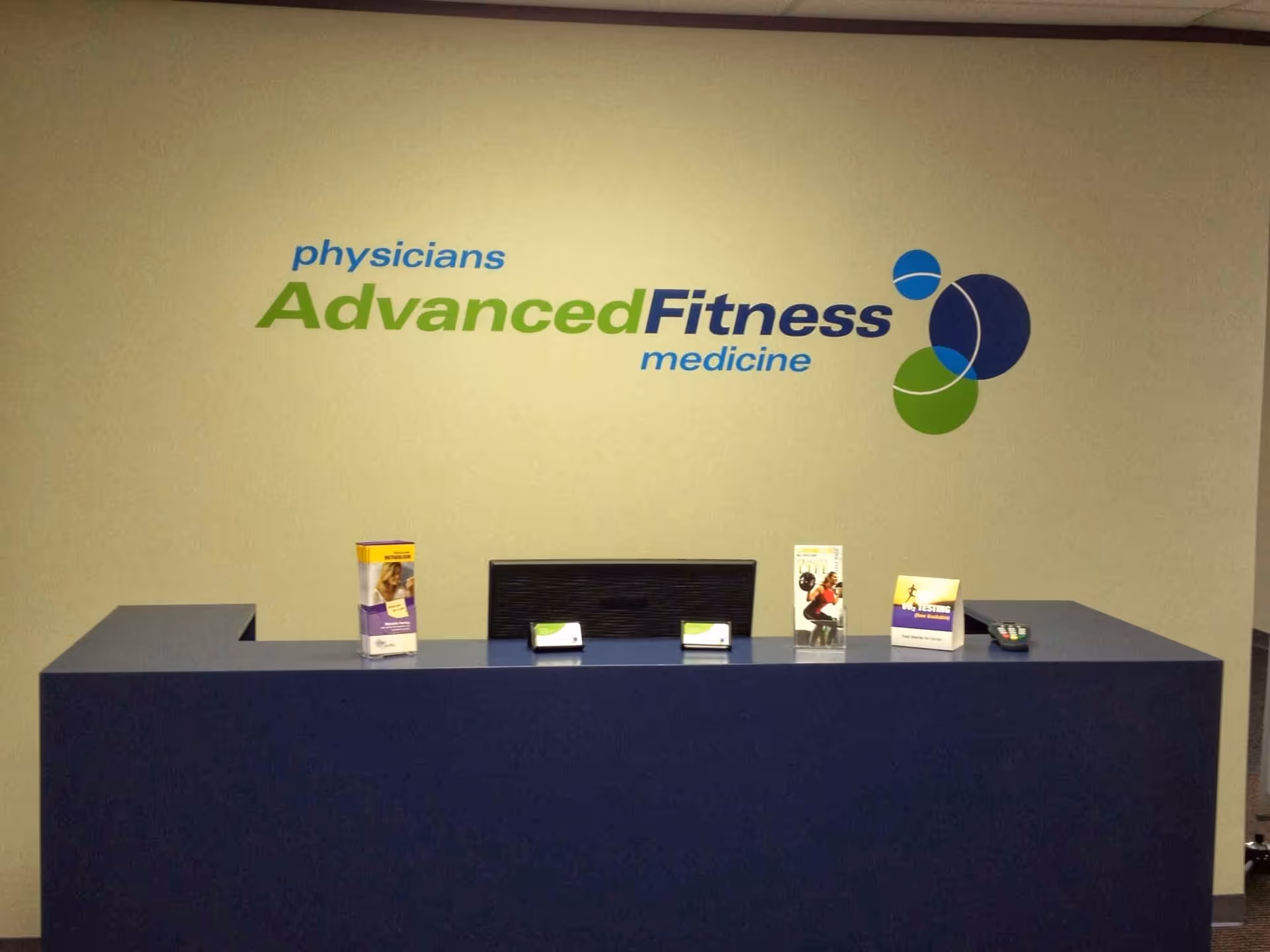Blue reception desk in a medical facility lobby with AdvancedFitness wall logo.