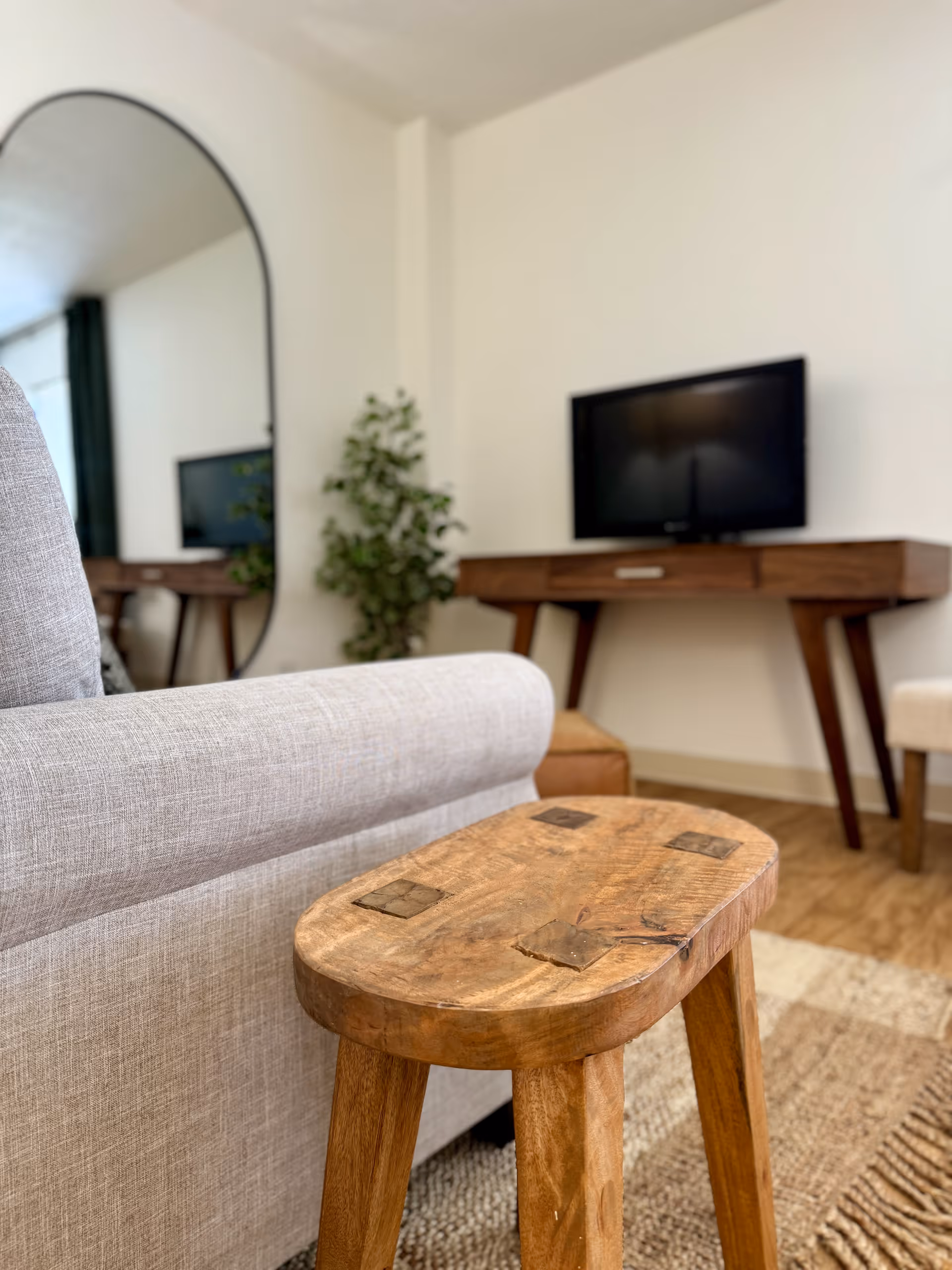 A cozy living room area featuring a light gray upholstered sofa, a rustic wooden stool in the foreground, a wooden console table with a flat-screen TV on top, a large oval mirror on the wall, and a green potted plant in the corner.