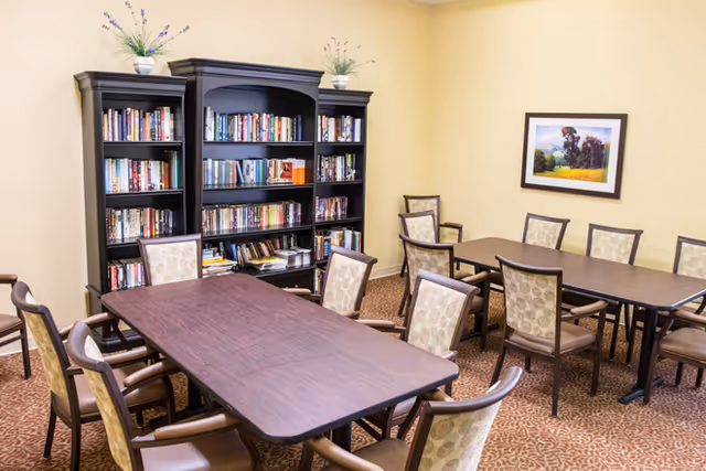 A cozy room with two rectangular wooden tables surrounded by cushioned chairs. Behind the tables is a large dark wooden bookshelf filled with books and decorated with two small potted plants on top. A framed landscape painting hangs on the beige wall. The floor is carpeted with a patterned design.