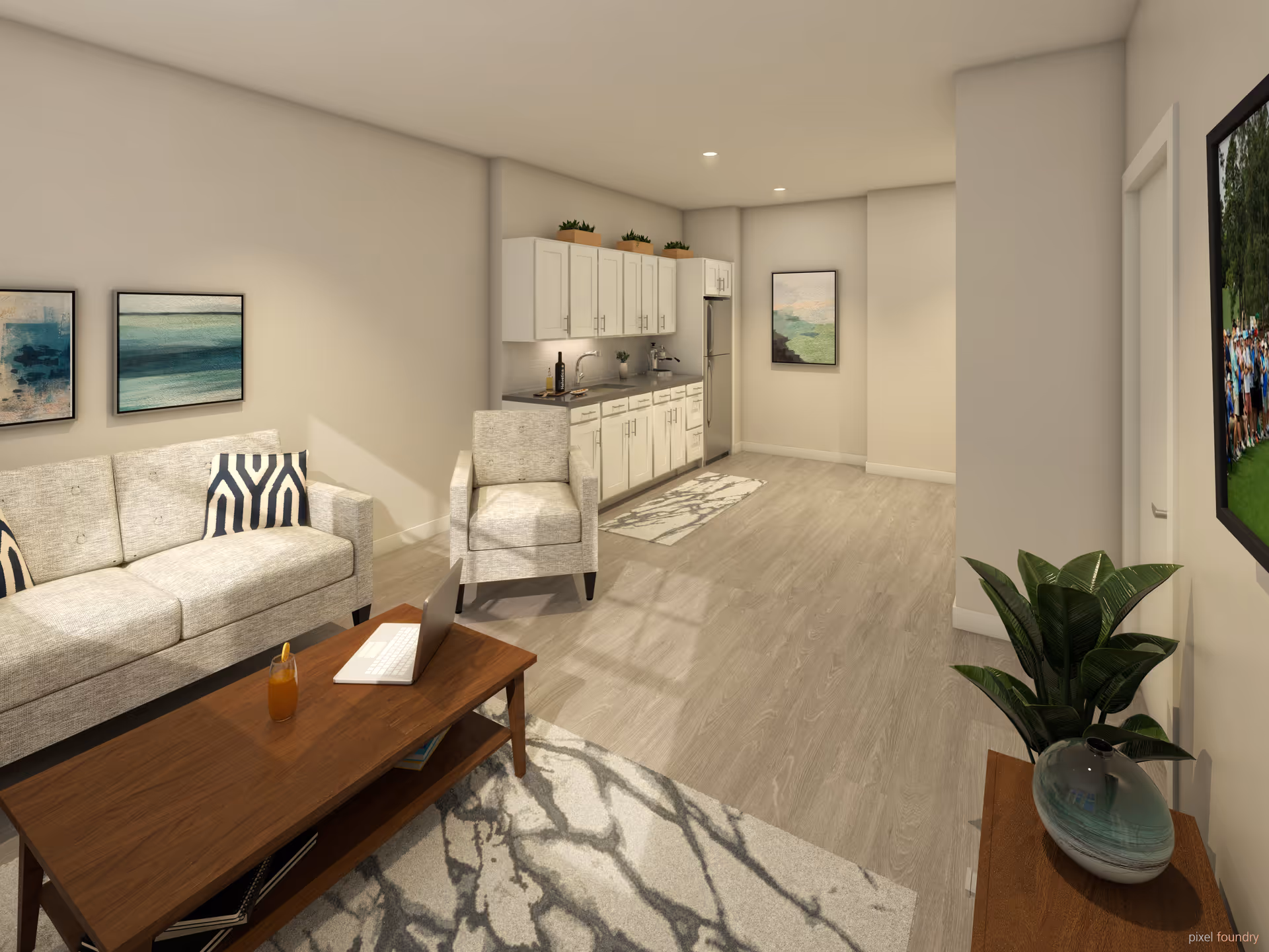A modern living room and kitchenette area with light-colored walls and flooring. The living room features a beige sofa with patterned pillows, a matching armchair, a wooden coffee table with a laptop and a drink on it, and a patterned rug. The kitchenette has white cabinets, a sink, a refrigerator, and small plants on top of the cabinets. There are framed artworks on the walls and a green plant on a wooden stand near the entrance.