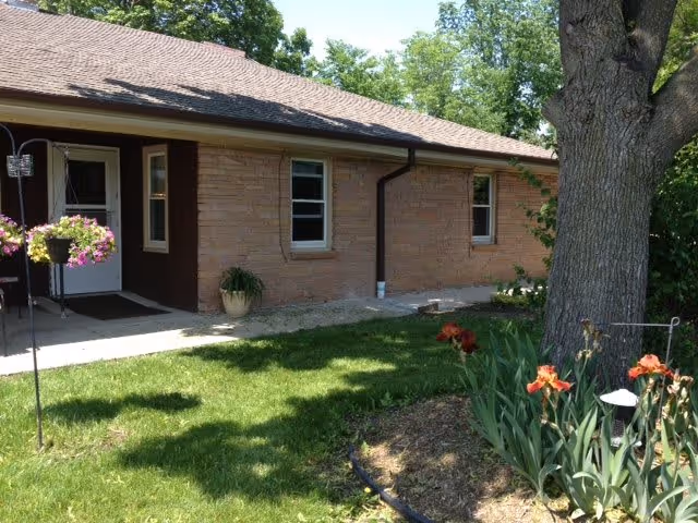 Exterior view of a single-story brick building with a brown shingled roof, two windows, and a white door. There is a green lawn with a flower bed containing orange flowers and a large tree in the foreground. A hanging flower basket is visible near the entrance.