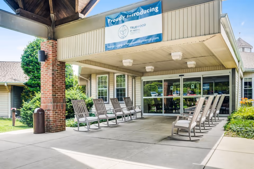 Covered entrance of Truewood by Merrill senior living with rows of outdoor rocking chairs under a porte-cochère and sliding glass doors.