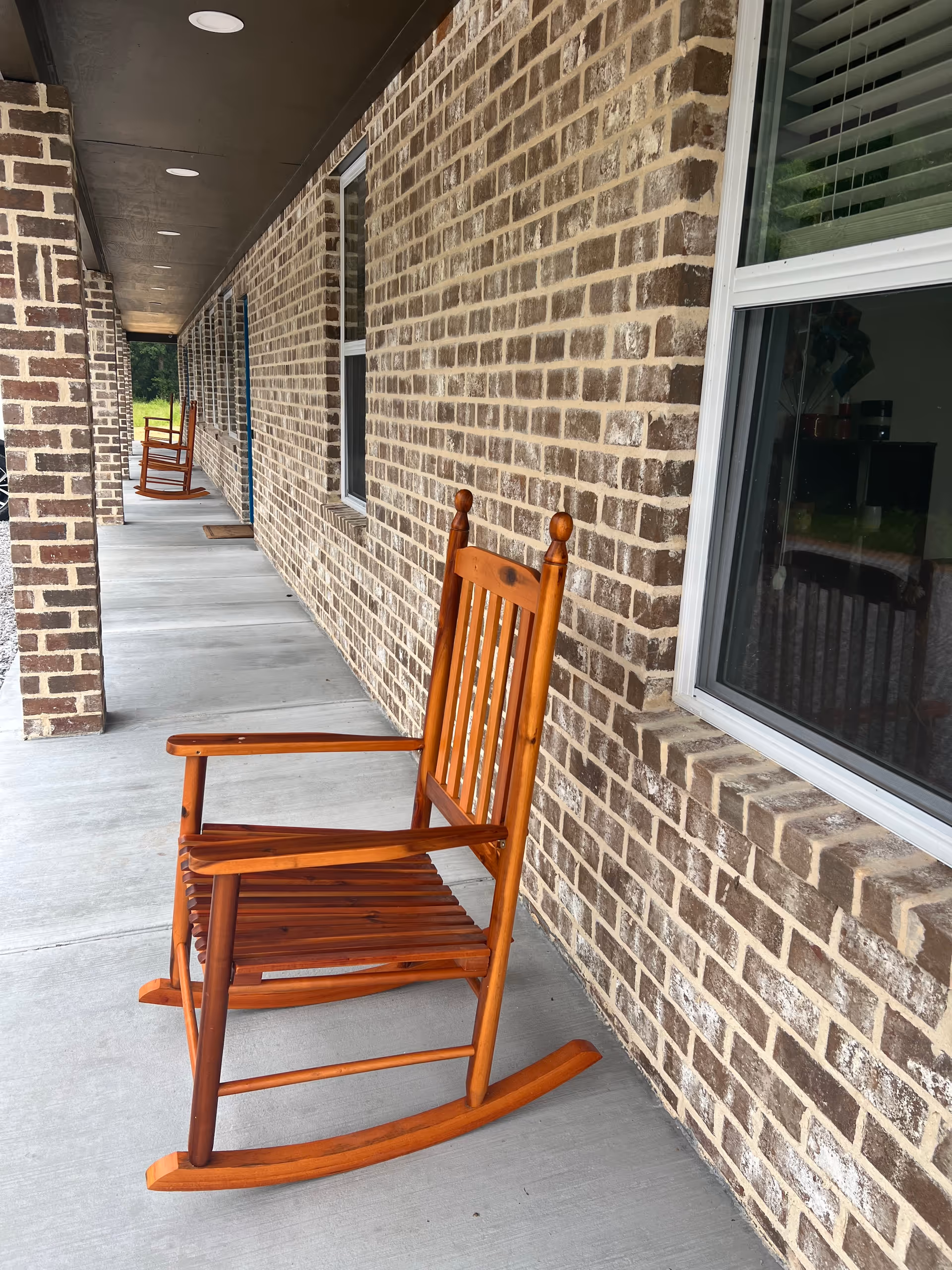 Row of wooden rocking chairs along a brick exterior walkway under a covered corridor.