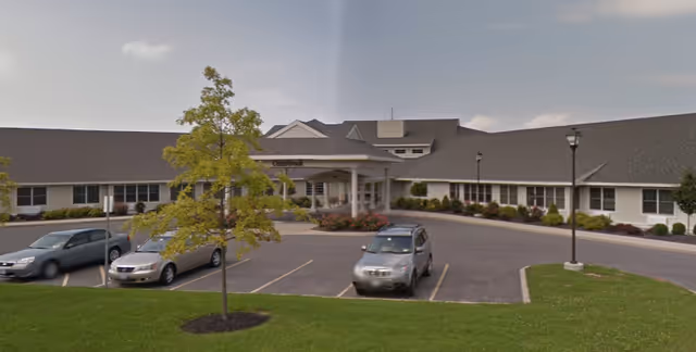 Exterior view of Cedarbrook senior living facility showing a large single-story building with a covered entrance, several parked cars, a tree, and a well-maintained lawn under a partly cloudy sky.