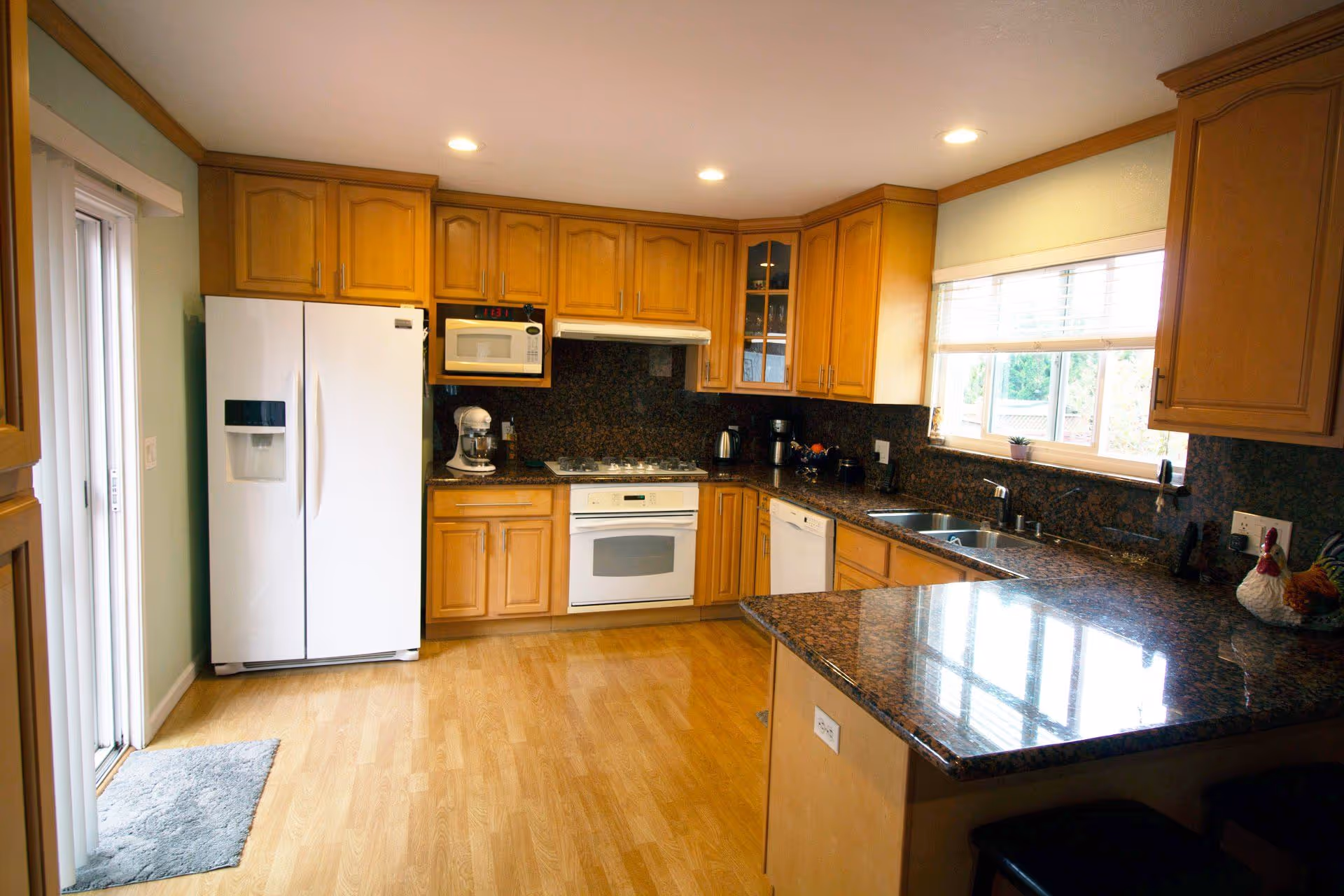 A bright kitchen with wooden cabinets, a white refrigerator, a microwave, a stove, a dishwasher, and a granite countertop. There is a window above the sink letting in natural light and a sliding glass door to the left with a small rug in front of it.