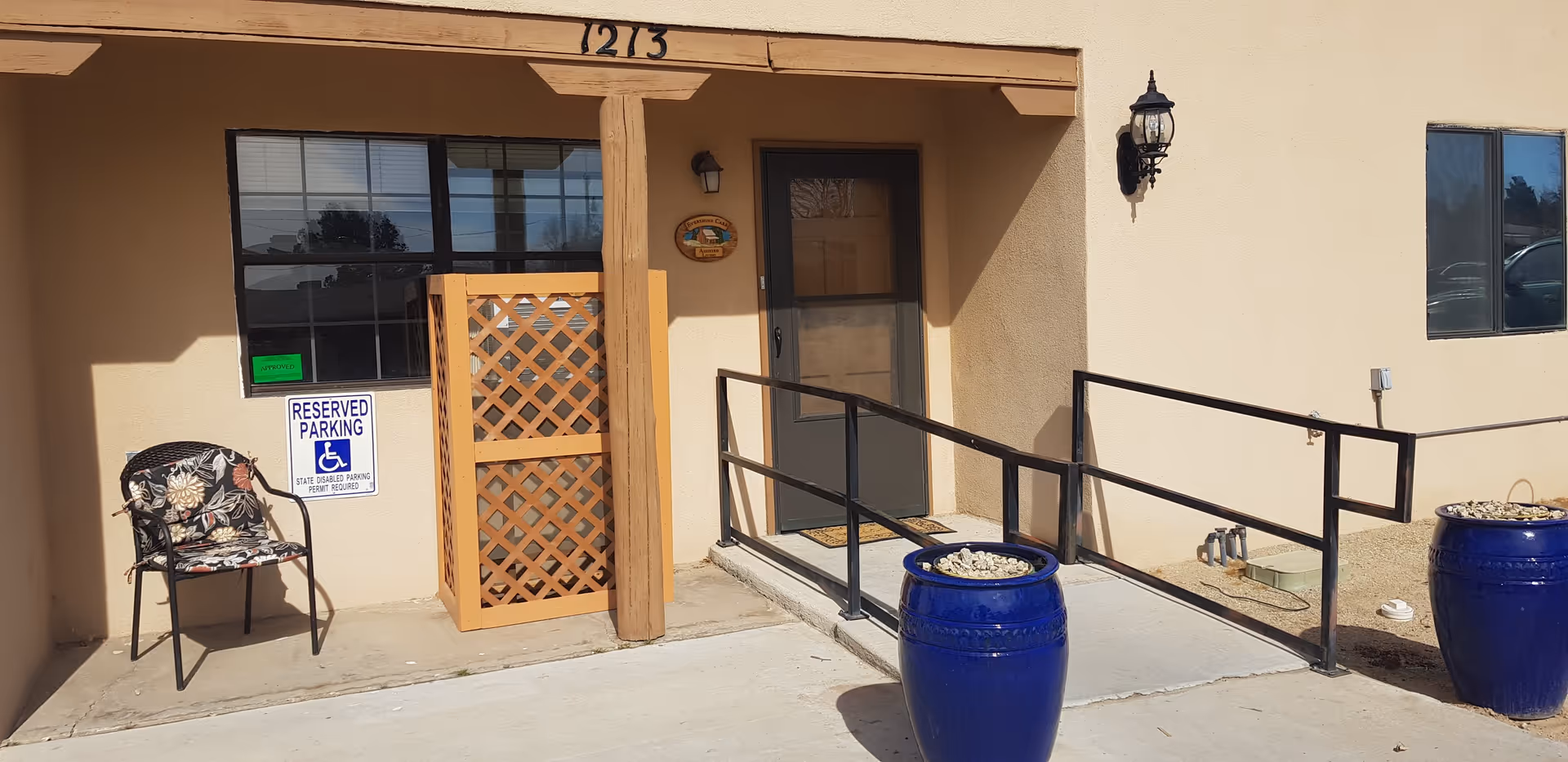 Entrance to a building with a ramp and handrails for accessibility. There is a door with a screen door, a window with a reserved parking sign for disabled parking, a chair with floral cushion, and two large blue planters filled with rocks. The building number 1213 is displayed above the entrance.