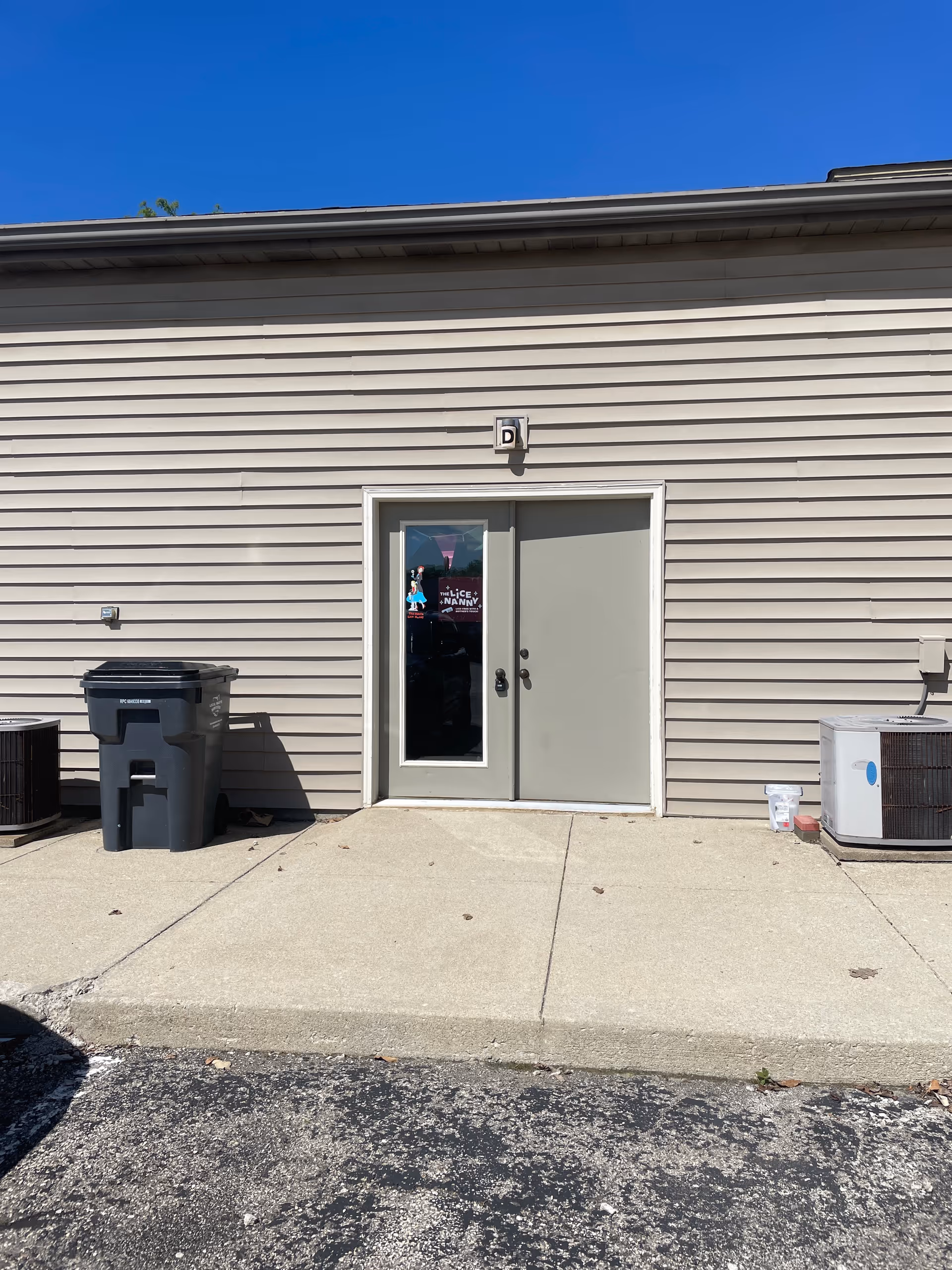 Exterior view of facility entrance with a glass door, beige siding, trash bin, and HVAC unit.