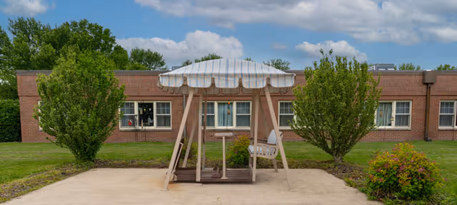 Outdoor courtyard with a striped canopy swing set, brick building, trees, and shrubbery.