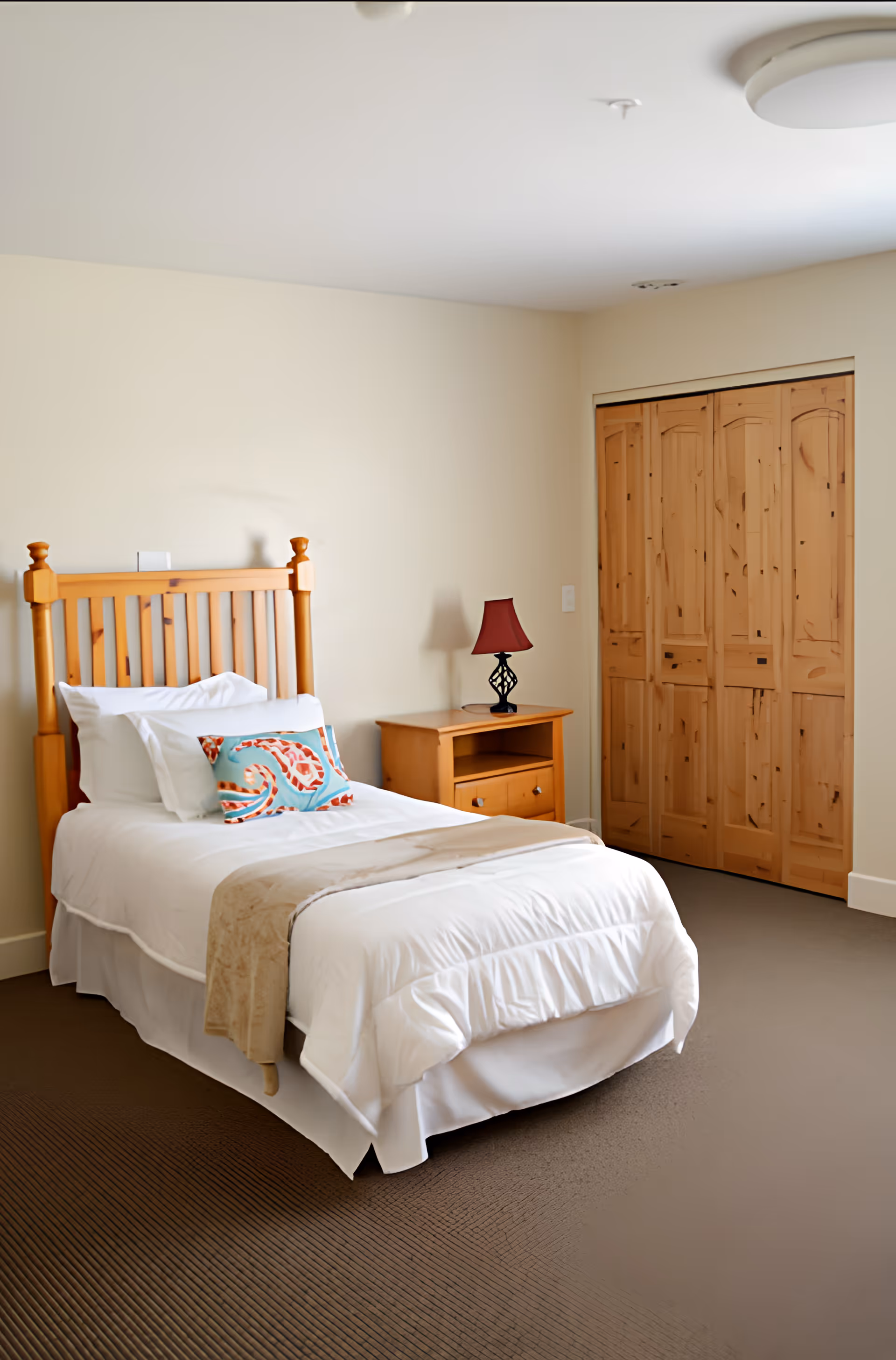 A tidy single bedroom with a wooden headboard, white bedding, a nightstand with a lamp, and closed wooden closet doors.