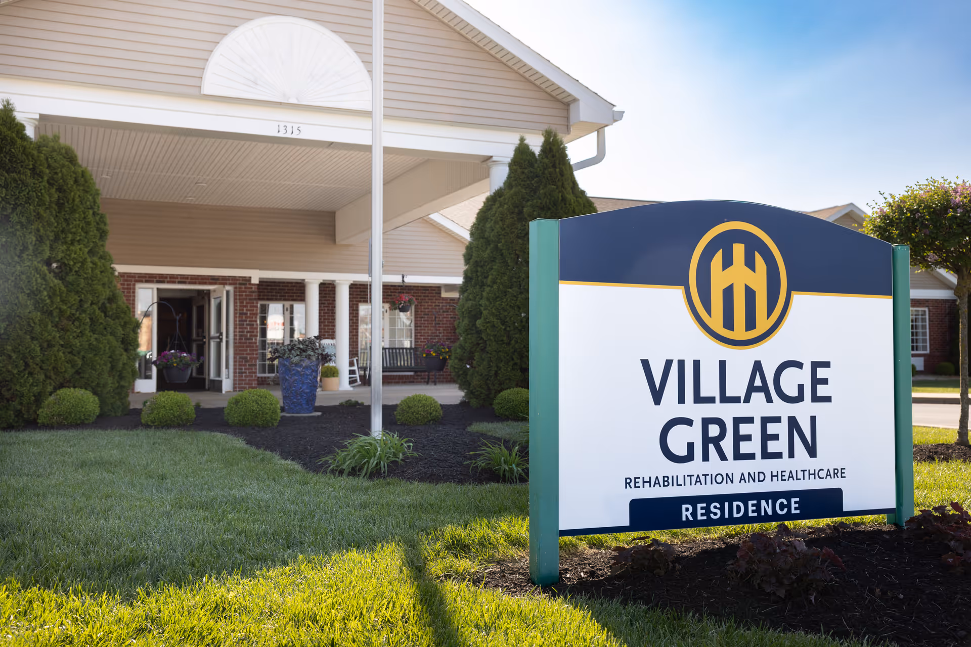 A Village Green sign sits near the front of a senior living building with landscaping.
