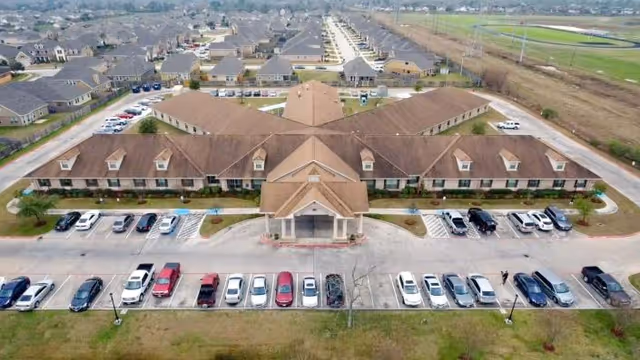 Aerial view of a single-story rehabilitation center with a central entrance, a parking lot of cars, and surrounding residential neighborhood.