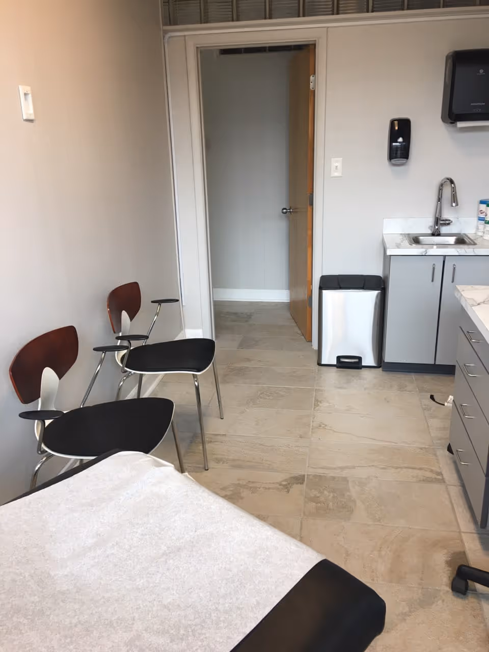 Clinical treatment room with a bed, three chairs, sink, and doorway.