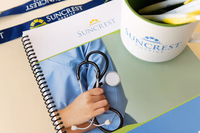 A hand holds a stethoscope over a notebook with Suncrest Hospice branding on a desk.