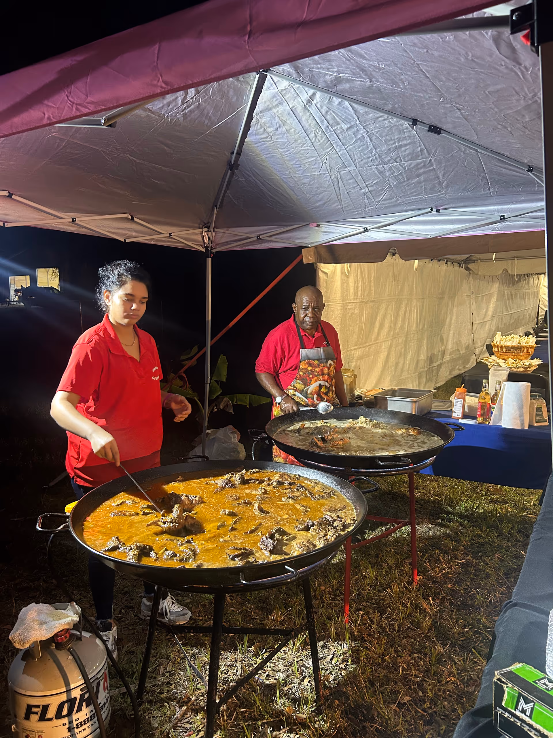 Two staff members cooking large pans under a canopy at an outdoor senior services event.