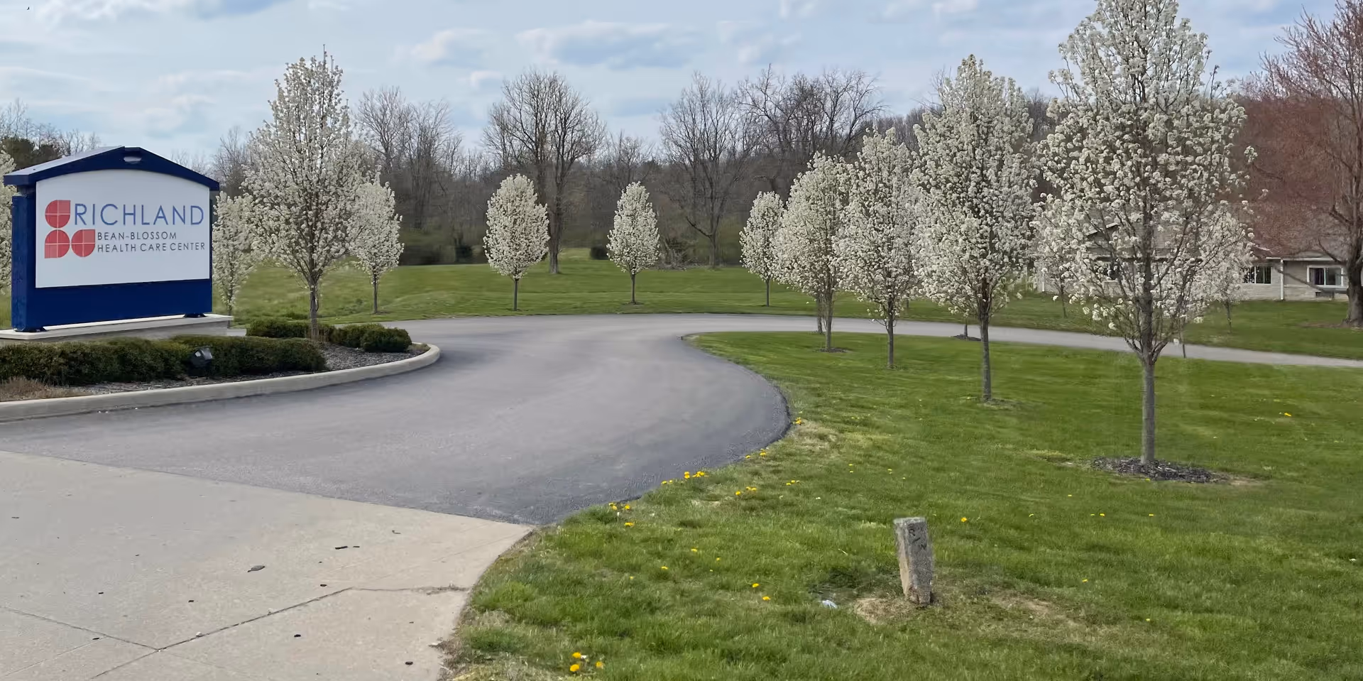 Blue-framed sign for Richland Bean-Blossom Health Care Center beside curved driveway and blossoming trees.