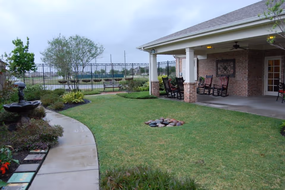 Well-kept outdoor courtyard with a covered patio and rocking chairs overlooking a lawn, walkway, fountain and fenced yard.