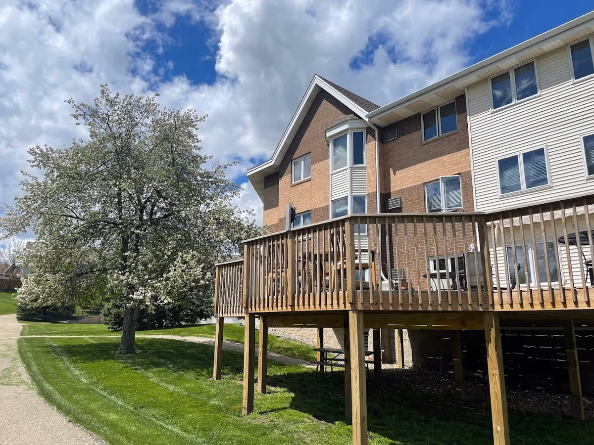 Multi-story brick-and-siding building with a raised wooden deck, green lawn and a flowering tree under a partly cloudy sky.