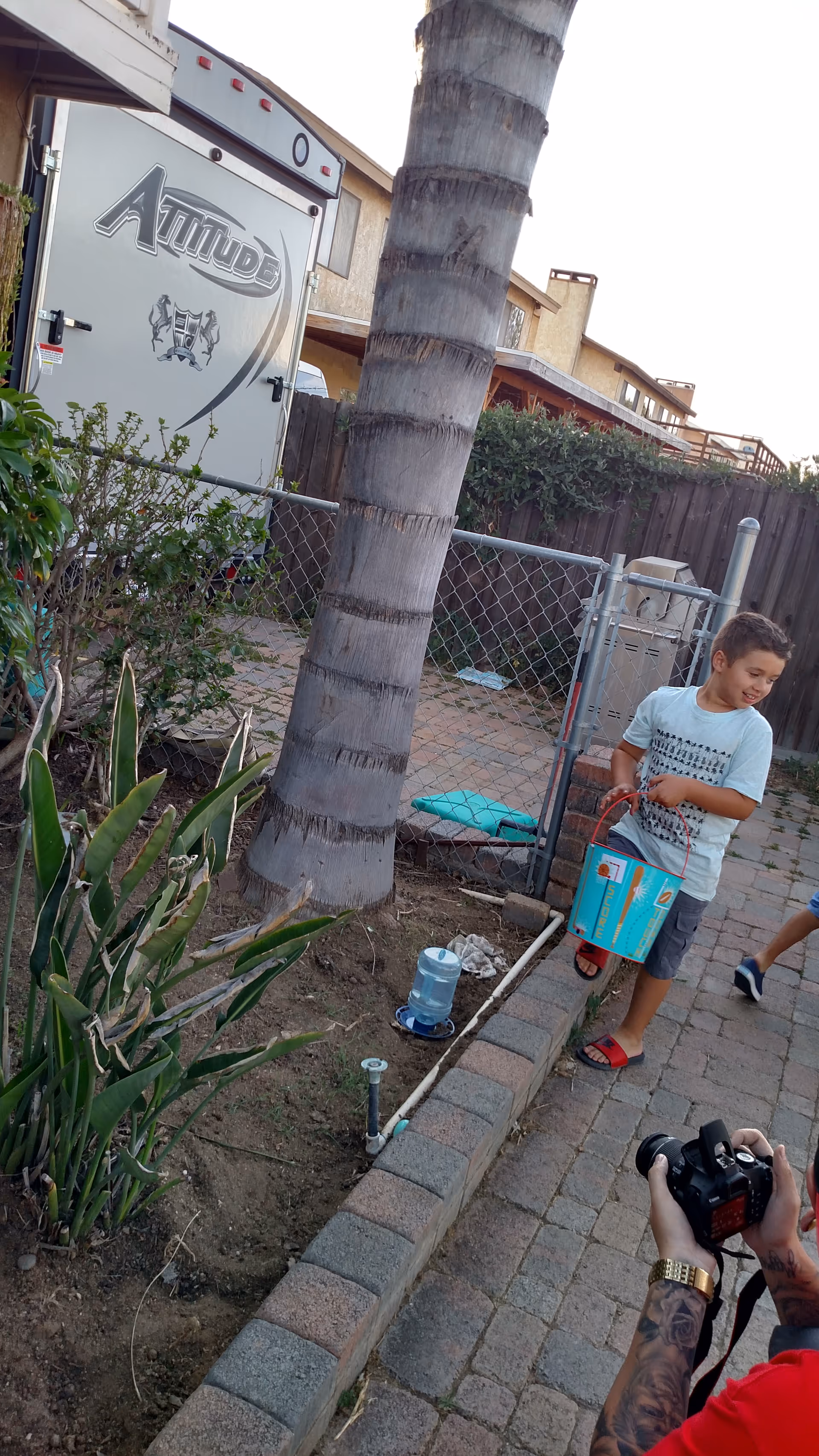 Outdoor courtyard with palm tree, fence, trailer, and a child holding a box beside a photographer.