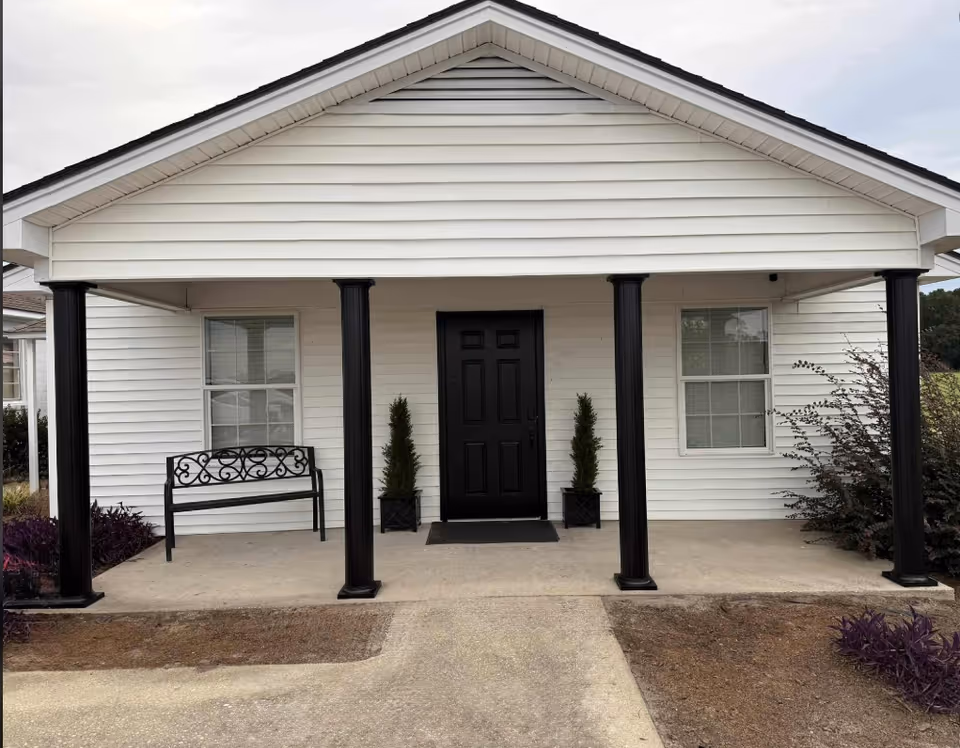 White single-story building front with a black door, columns, porch, bench, and potted shrubs