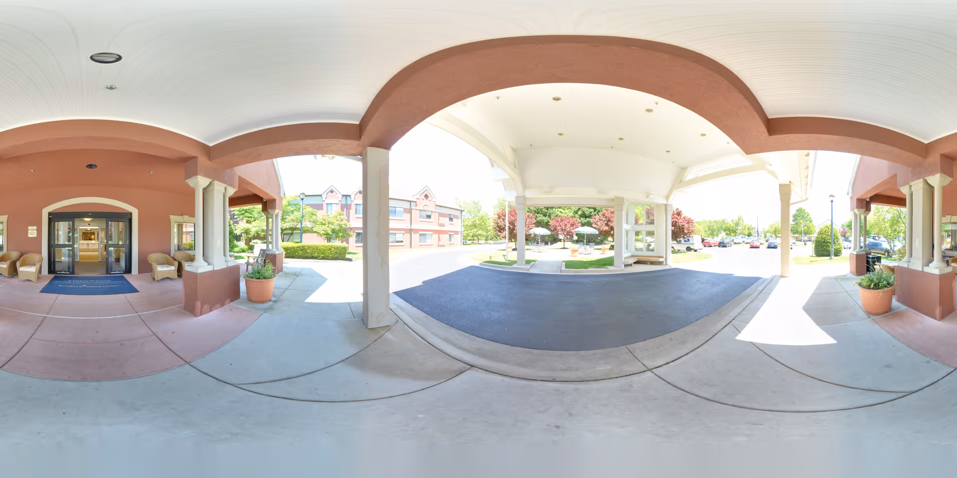 Covered entrance area of Aspenwood Senior Living Community with a driveway for vehicles, potted plants, seating chairs, and a view of the building and parking lot in the background under a bright sky.