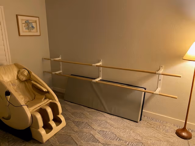 A small therapy room with a massage chair, wall barre, folded mat, and floor lamp.