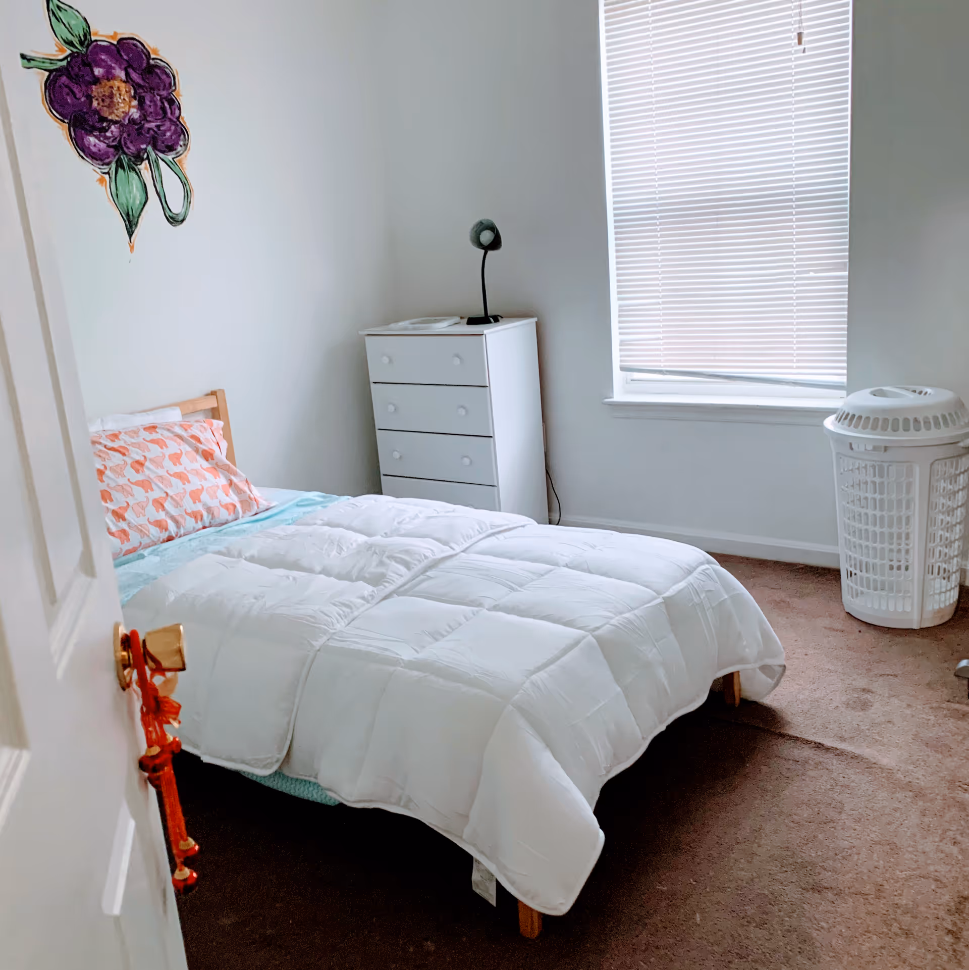 A small bedroom with a single bed covered in a white comforter and a pillow with a flamingo pattern. There is a white dresser with a small black lamp on top, a window with closed blinds, a white laundry basket, and a purple flower decoration on the wall.
