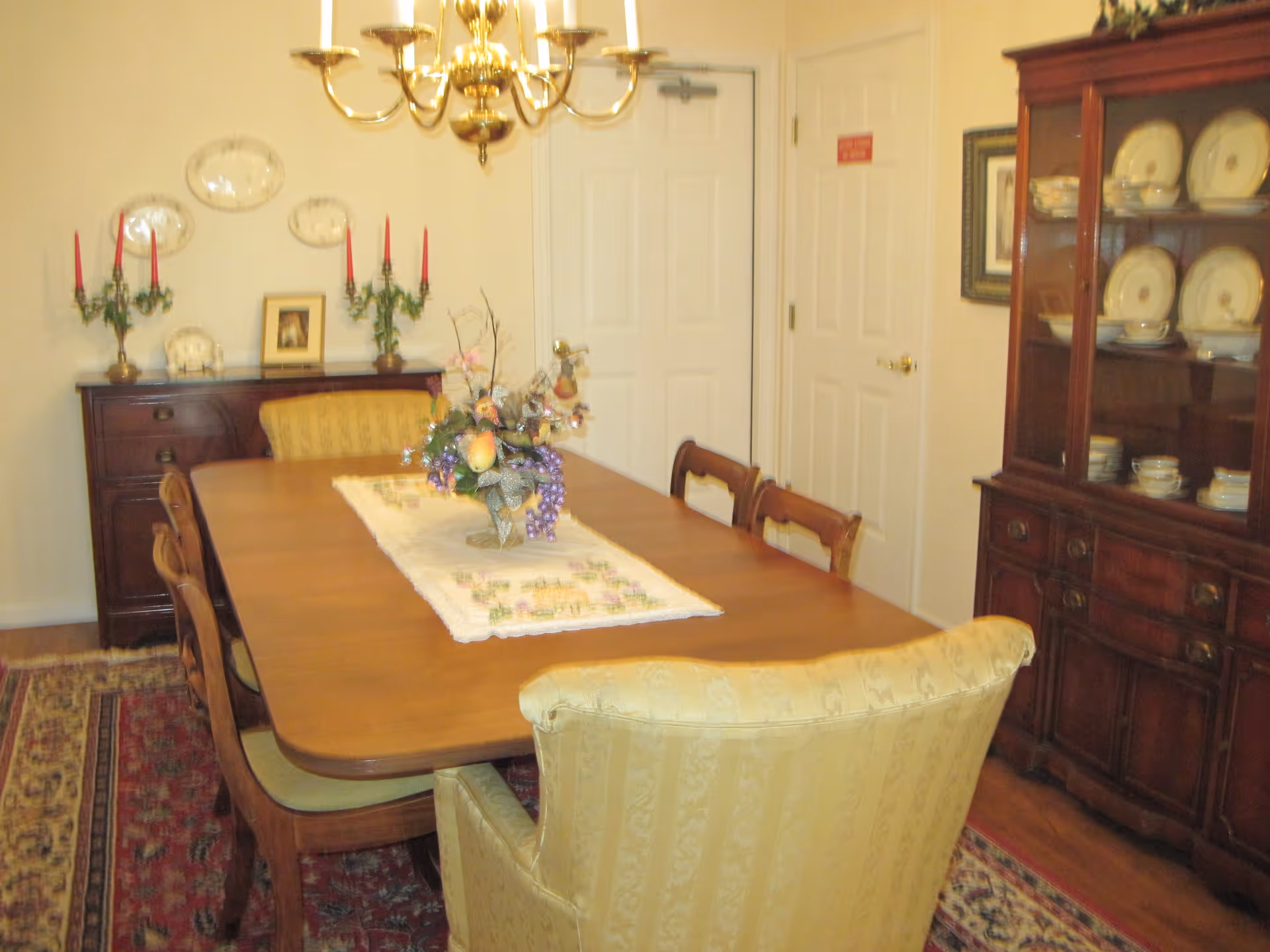 A traditional dining room with a wooden dining table surrounded by six chairs, including two upholstered armchairs at the ends. The table has a decorative runner and a floral centerpiece. On the right side, there is a wooden china cabinet displaying plates and bowls. On the left side, there is a sideboard with two candelabras holding red candles, a framed picture, and decorative plates mounted on the wall above. A brass chandelier hangs above the table, and a patterned rug covers the floor.
