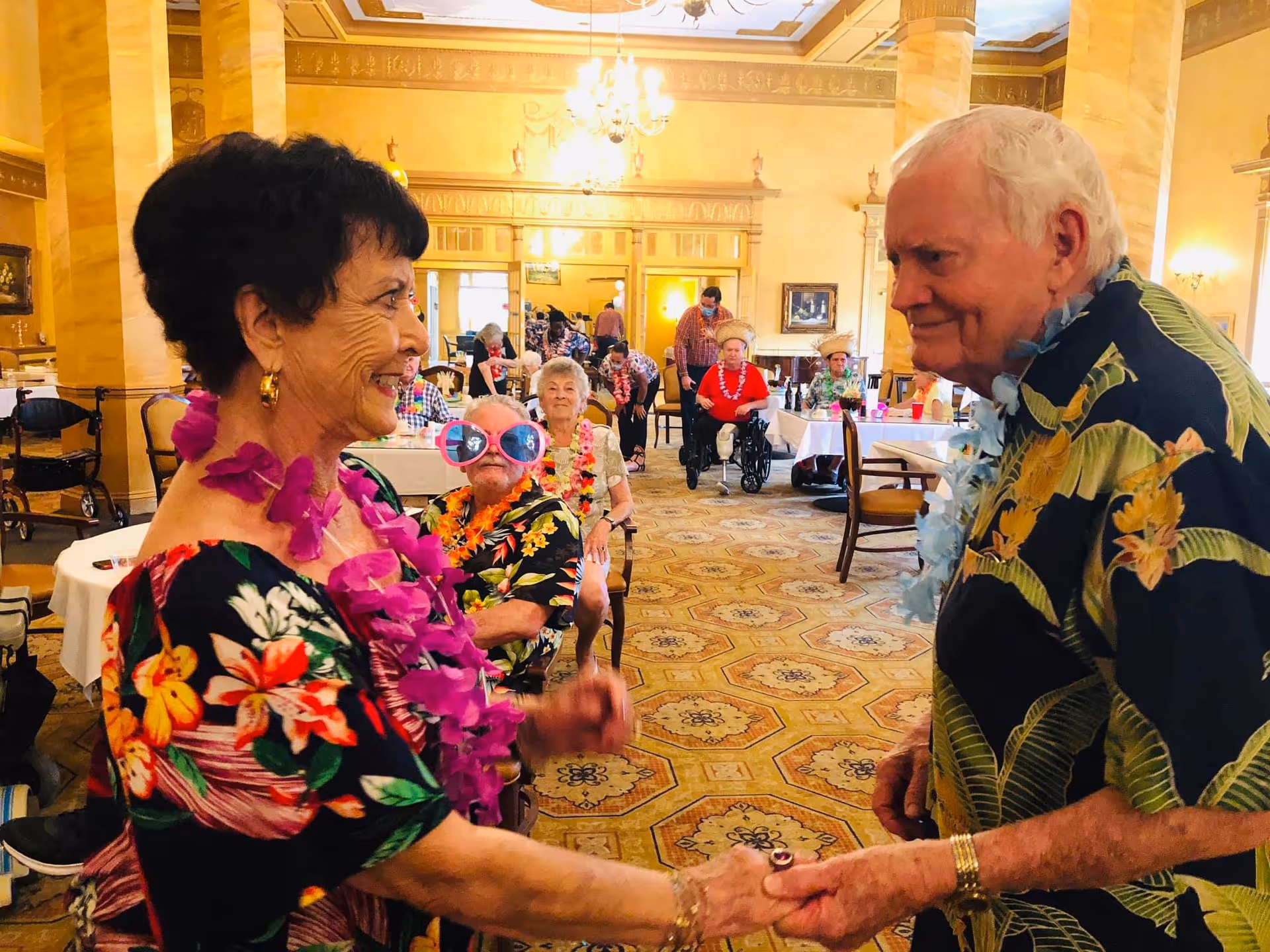 Two elderly residents in Hawaiian shirts and leis holding hands and smiling in a decorated dining room with other seniors seated at tables.
