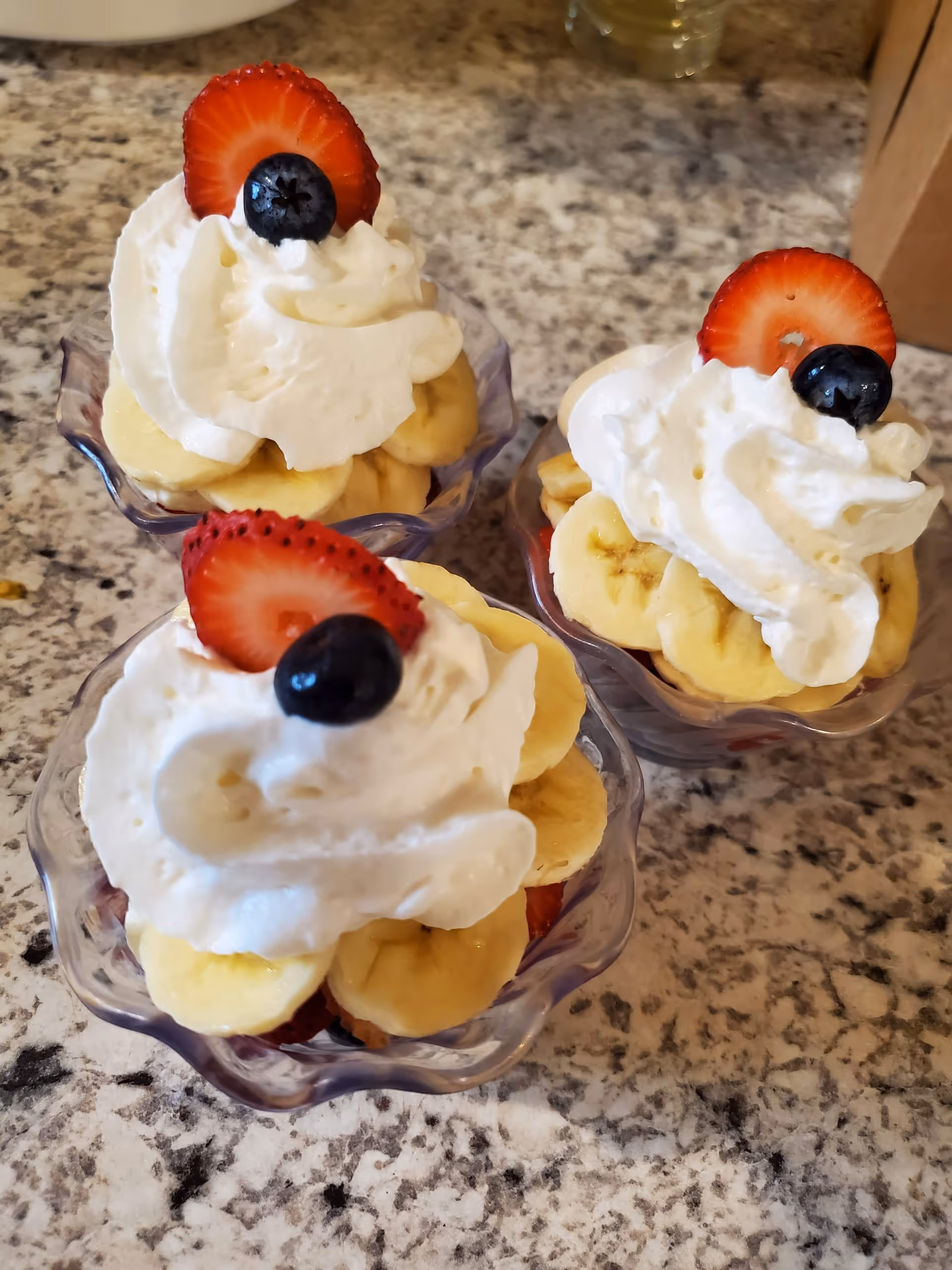 Three glass dessert cups topped with bananas, strawberries, blueberries, and whipped cream on a granite counter.