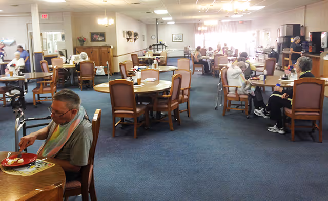 A dining room in a senior living facility with several elderly residents seated at round tables, eating and conversing. The room has blue carpet, wooden chairs with cushions, and soft lighting. There is a kitchen area visible in the background with appliances and a counter.