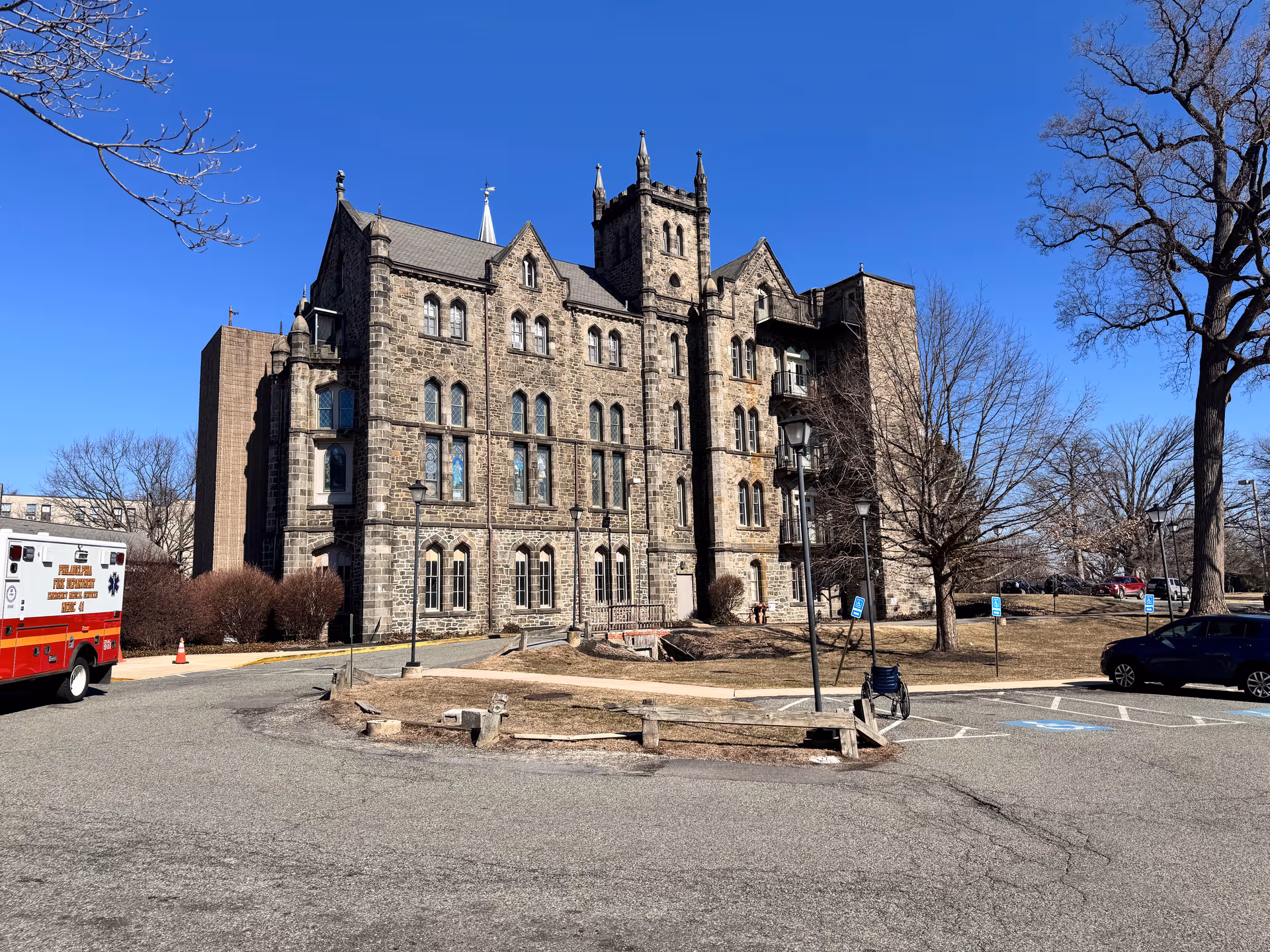Stone Gothic-style senior care building with parking lot, ambulance, and leafless trees on a clear day.