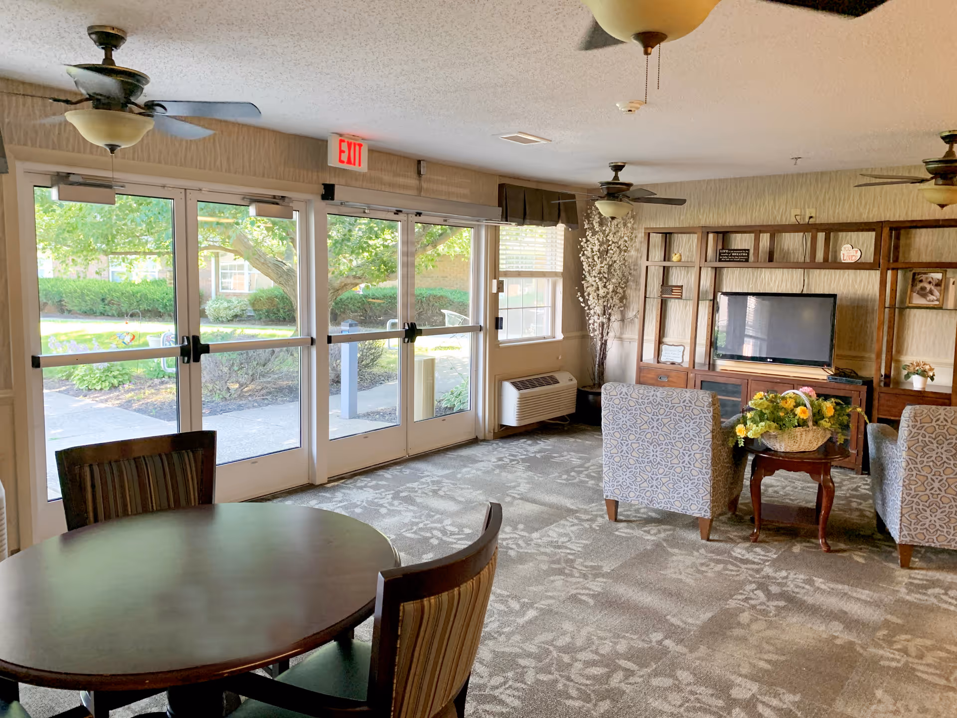 A bright and cozy common area in a senior living facility with large glass doors and windows letting in natural light. The room features a round wooden table with chairs, two patterned armchairs facing a flat-screen TV on a wooden entertainment center, ceiling fans, and a small table with a flower basket. Outside, greenery and a walkway are visible.