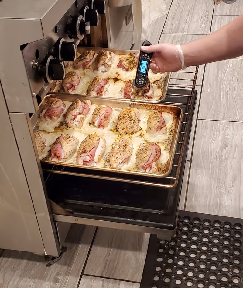 Chicken portions bake in metal trays as staff checks temperature with a digital thermometer.