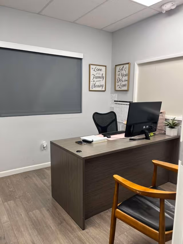 An office desk with computer, chair, and framed wall decor in a senior living facility.