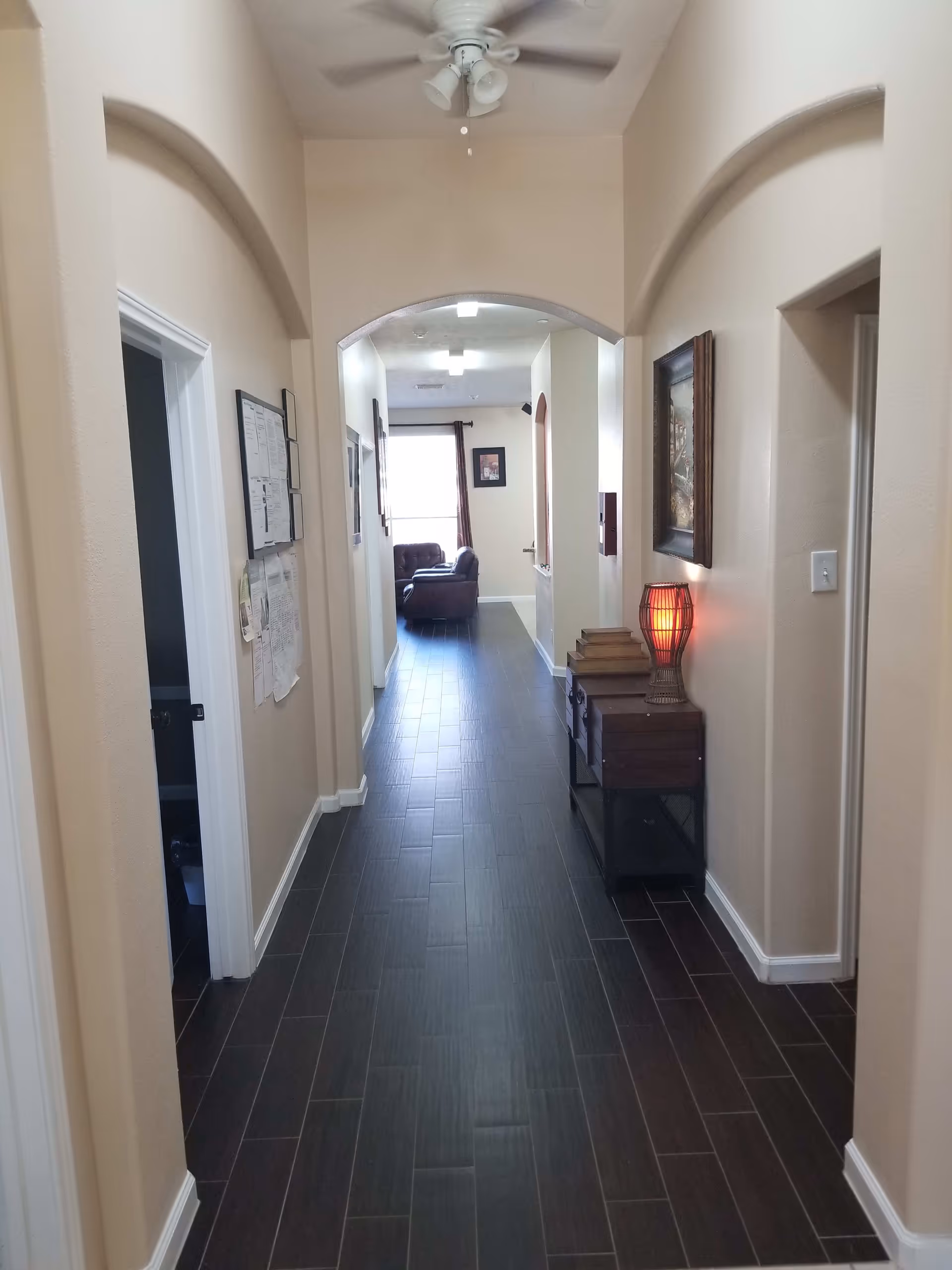 Long interior hallway with dark wood-look tile flooring, arched doorways, a small console table with a lit lamp, and seating visible at the far end.