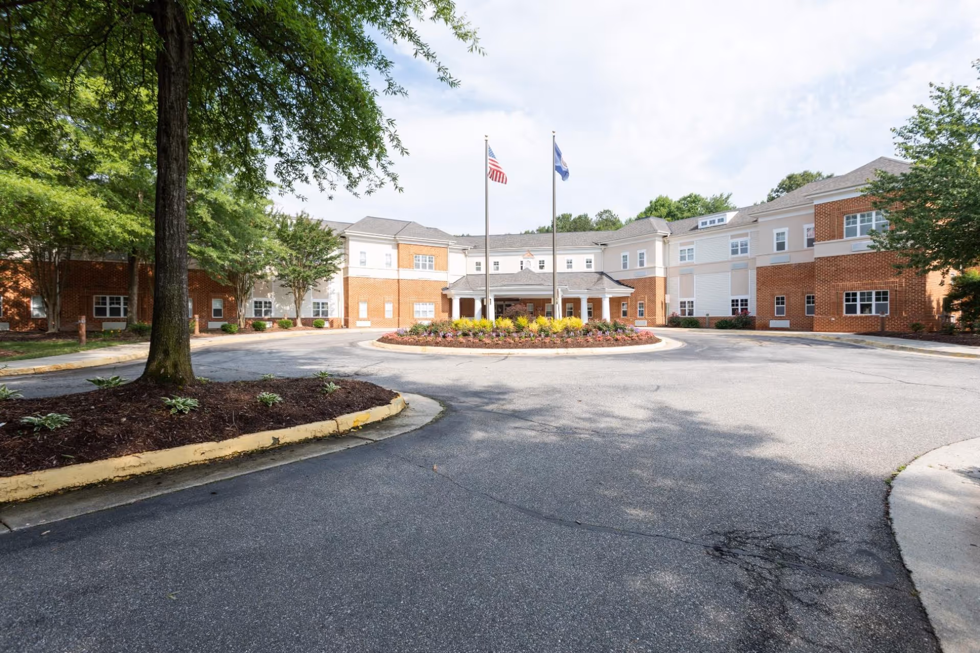 Front exterior view of a large two-story senior living facility building with a circular driveway and landscaped roundabout featuring flowers and two flagpoles with the American flag and another flag. Trees and greenery surround the building under a partly cloudy sky.